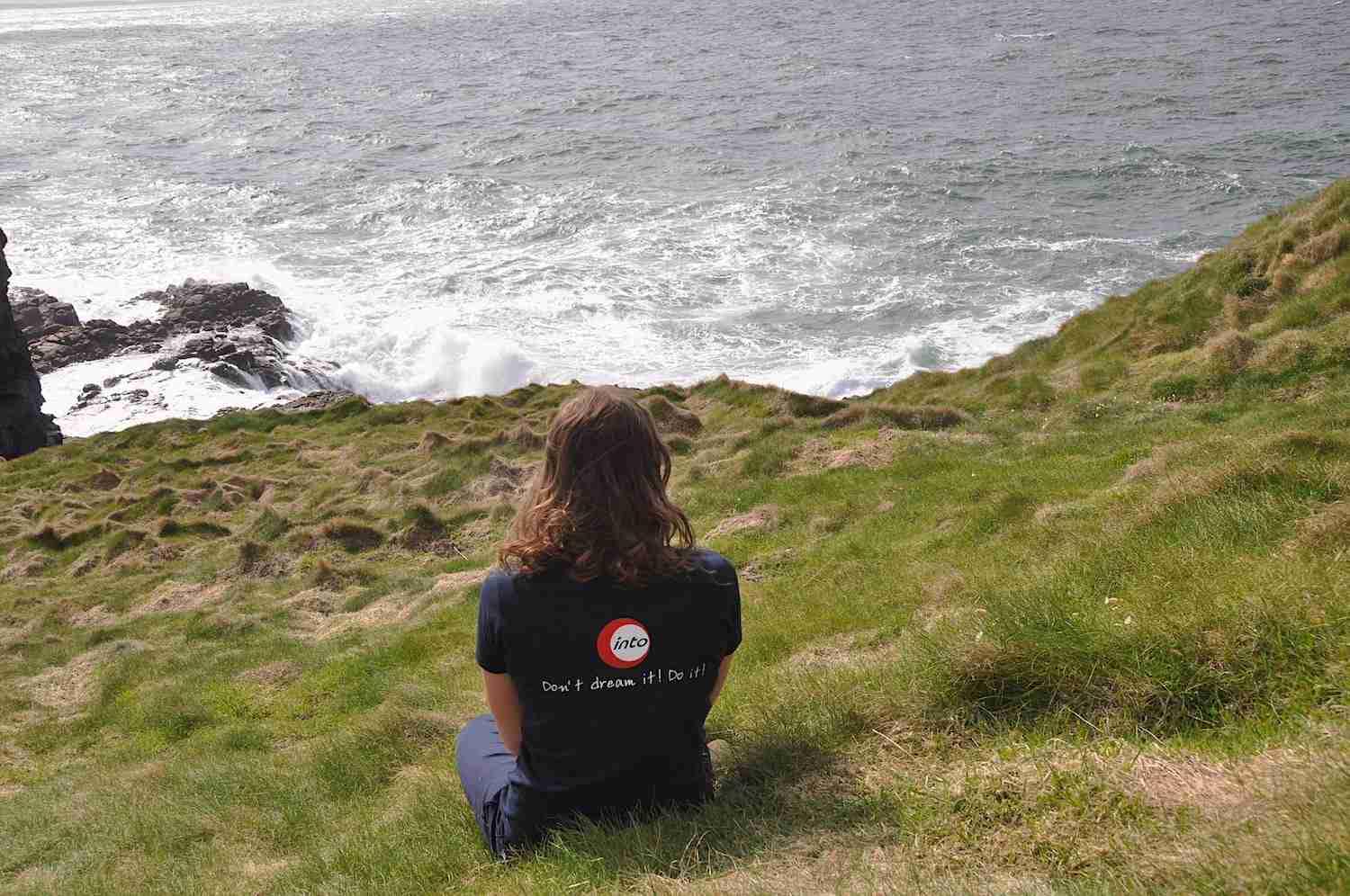 Into uitwisseling student in Ierland Achterzijde van een meisje zittend op een klif en kijkend naar de zee met een blauw T'shirt met into logo.